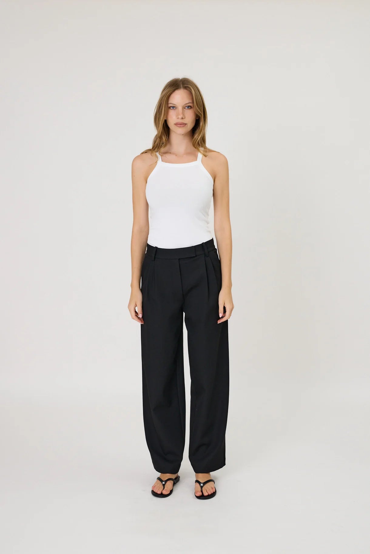 Woman wearing Remain Lindy black wide-leg pleated suiting trousers with black thong sandals.